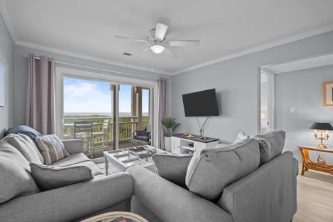 Oak Island, NC living room with gray sofas, wall TV, ceiling fan, sliding doors, ocean-view deck, coastal décor, and bright beach house style.