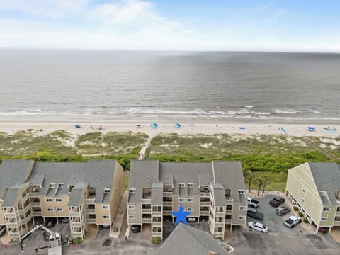 Aerial view of Oak Island, NC condos with blue star marker, parking, balconies, dunes, beach access, umbrellas, sandy shoreline, waves, and wide Atlantic Ocean views.