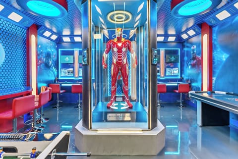 Welcome to a lively entertainment room where bold blue and red tones energize the space. Enjoy the stunning suit in the futuristic display case while relaxing on sleek bar seating, perfect for socializing and having a great time.