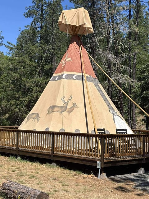 26-foot authentic tipi on an oversized deck with cedar railing—outdoor rocking chairs.  Towering lodgepoles rise 30 feet overhead, draped in hand-painted canvas that glows amber at sunrise and flickers gold by the firelight at night.  