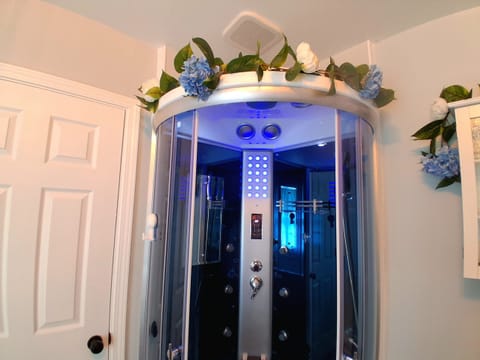 Enter your own spa sanctuary featuring a combination steam‑sauna and rainfall‑shower unit illuminated by calming blue lighting. Across the room, a heated two‑person jetted tub creates a soothing, restorative atmosphere.
