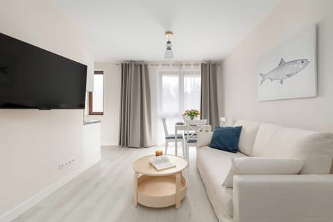 A bright living room with a white sofa, a wall-mounted TV, and a small round coffee table placed on light wood flooring.