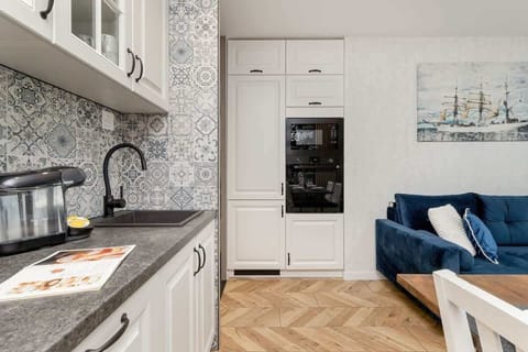 A bright kitchen with white cabinetry, wooden countertop, and black-and-white patterned backsplash. A living space is visible beyond the kitchen.