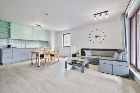 A bright, modern living room with a gray couch, a glass coffee table, and a dining area in the background. The room is well-lit and features contemporary decor.