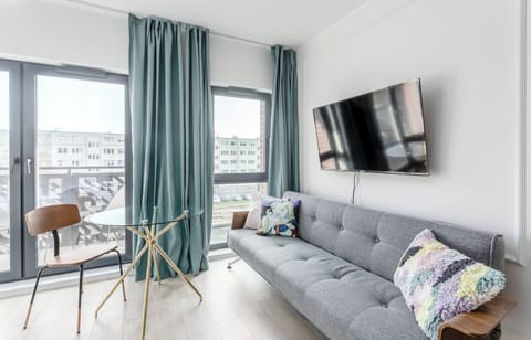 A bright living room with a sofa, TV, and small dining area by a large window. The modern design creates a cozy atmosphere with natural light.