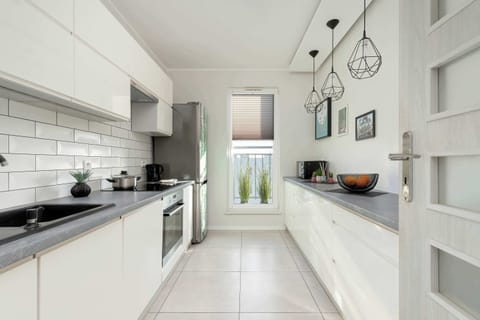 A sleek, white kitchen with modern cabinetry, a black countertop, and pendant lighting. The space is well-organized and features a built-in oven and a large window for natural light.