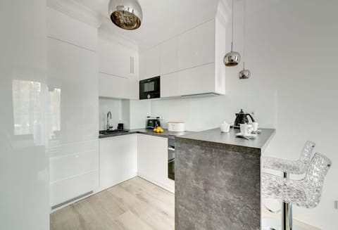 A sleek, modern kitchen with glossy white cabinetry, a minimalist design, a small black sink, built-in appliances, and a gray breakfast bar paired with transparent bar stools, complemented by metallic pendant lights.