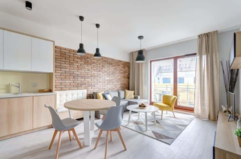 A cozy living area with a stylish mix of neutral tones, a modern round dining table, a comfortable gray sofa with colorful cushions, and a yellow accent chair, complemented by a brick wall and large windows that let in natural light.