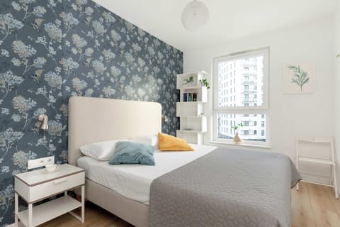 A bedroom with a neatly made bed featuring white and yellow pillows, a wooden bedside table, and a stylish blue-patterned accent wall.