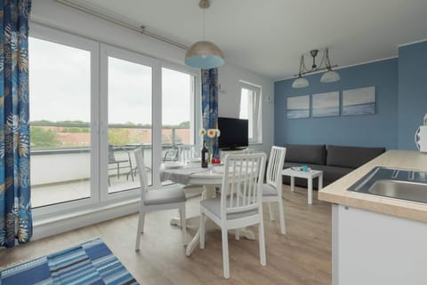 A bright, spacious living area with large windows, a small dining table, and modern decor. The room has blue walls, giving it a calm, coastal feel.
