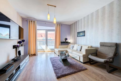 A stylish living space featuring light wooden flooring, a modern gray sectional sofa paired with matching armchairs, and a sleek, wall-mounted flat-screen TV. A decorative light fixture hangs from the ceiling, creating a warm and inviting atmosphere.