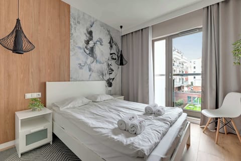 A stylish bedroom with a large bed, white bedding, and a unique artistic mural on the wall. Floor-to-ceiling curtains frame a bright window, allowing natural light to fill the room.