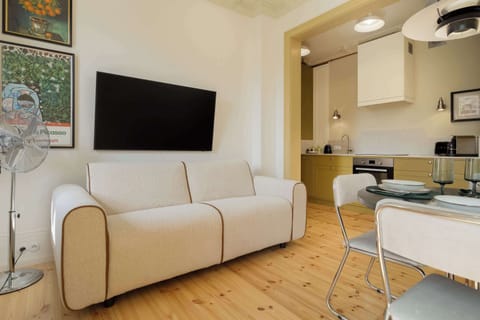 An open-plan living room with a small kitchen visible behind a cozy seating area and a wall-mounted TV.