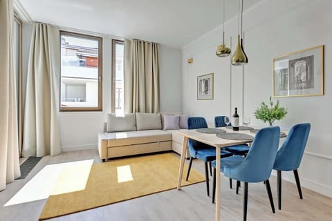 A well-lit living room with a modern sofa, a small coffee table, and dining chairs beside a large window.
