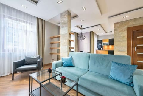 A modern living area with a vibrant teal sofa, wooden elements, and natural light streaming through large windows.
