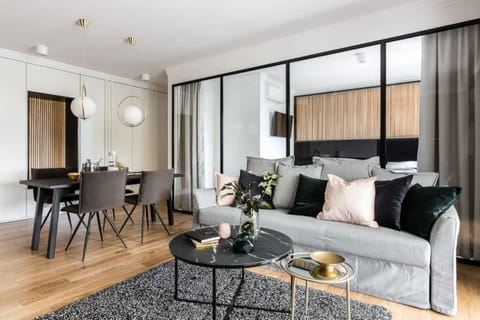 A modern living room with a stylish grey couch and a large black coffee table. The space flows into a dining area with a sleek round table and four chairs. Natural light fills the room through large windows.
