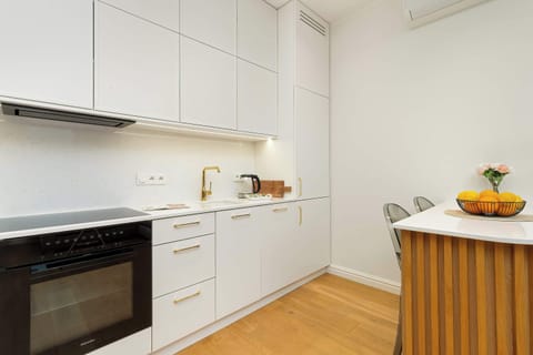 In the photo, we see a bright, modern kitchen with white cabinets, gold handles and a light-coloured worktop that connects to a dining area with a table and chairs.
