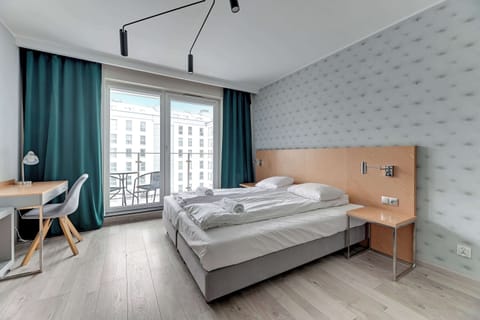 Cozy bedroom with a double bed covered in white linens. The room features light walls and a patterned blue accent wall, creating a soft and inviting atmosphere. Large windows let in ample natural light.
