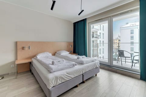 A bright living area with a double bed covered in white linens. A large window provides natural light and a view, while the modern decor includes light wood and soft colors.