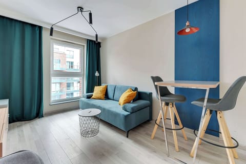 Cozy living area, a teal sofa is paired with a small round coffee table and a modern floor lamp. Large windows with green curtains allow natural light to brighten the space.
