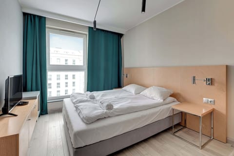 Modern studio with a queen-size bed and white linens. The room features a beige wall and a large window with curtains, providing natural light and a cozy atmosphere.