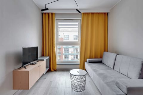 In the living area, there is a gray sofa paired with a small round white table. Bright yellow curtains frame a large window, and a TV is positioned on a wooden cabinet, creating a comfortable and inviting space.