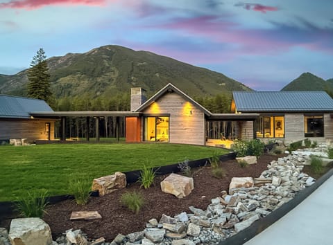 Modern mountain retreat with warm exterior lighting, custom landscaping, and stunning views of the surrounding peaks at sunset.