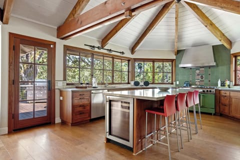 Bright open-concept kitchen and dining area with vaulted wooden beams, a large island with red bar stools, and extensive windows providing natural light.