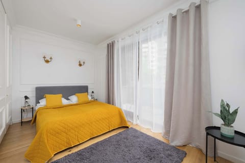 A bright bedroom area with a yellow bedspread, small sofa, and floor-to-ceiling curtains letting in natural light.
