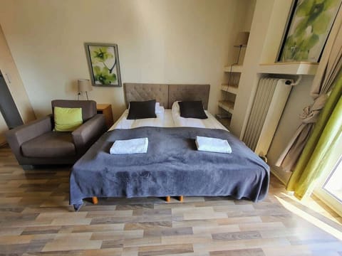 This cozy bedroom features a queen-sized bed with a blue comforter and pillows. There are bedside tables with lamps, a potted plant near the window, and a warm, inviting ambiance created by soft natural lighting.