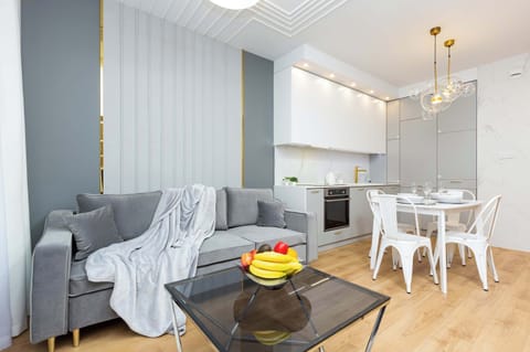 A modern living space featuring a grey sofa, a dining area with a white table and chairs, and a fully equipped kitchen. The room is open, airy, and tastefully decorated with neutral tones.