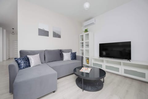 A cozy living room with a large gray sofa, a round coffee table, and a TV on a white media unit. The room is bright with sheer curtains letting in natural light.