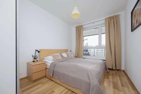 A cozy bedroom with a large bed, a wooden nightstand, and a window letting in natural light. The space is minimal and inviting, with a soft neutral color palette.