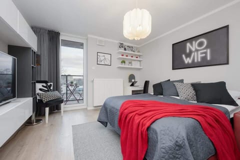 A modern studio apartment with a stylish bed dressed in red and gray, a "No WiFi" artwork on the wall, and a large window leading to a balcony.

