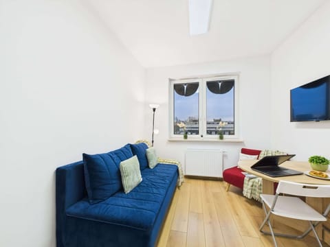 A cozy living room space features a blue sofa and a small dining area. The room is bright, with a wooden floor and large windows providing natural light.