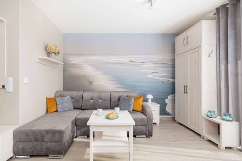 Modern living room with a sectional sofa, gray and orange pillows, and a large scenic wall mural depicting a seaside view. There is a coffee table and large windows letting in plenty of natural light.