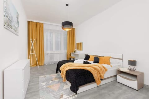 A bright bedroom with a comfortable double bed, yellow accents, and minimalist decor. Natural light fills the room through a large window with mustard curtains.