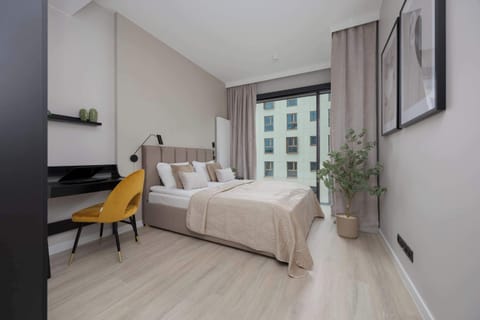 A cozy bedroom with a double bed, a workspace with a yellow chair, and large windows letting in plenty of natural light.