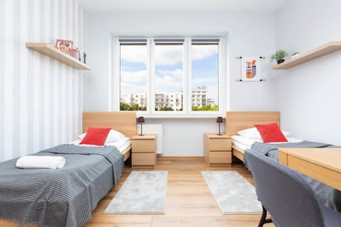 A room with two twin beds featuring gray bedding and red accents. The large window allows plenty of natural light, illuminating the wooden floor and simple decor.