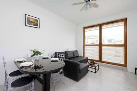 The bright living area features a sofa and a dining table, along with large windows that brighten the space with natural light.
