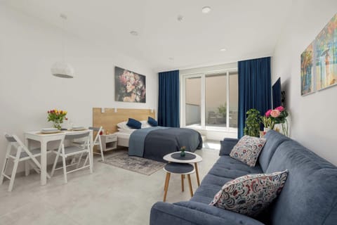 A spacious living area with blue sofas arranged around a small dining table. Bright white walls and soft lighting make the room feel open. Decorative artwork adds color and personality.