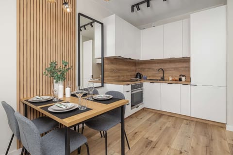 A compact kitchen combining white cabinetry with a wooden backsplash. A small dining table with two chairs stands nearby, complemented by a wood-accented wall panel.

