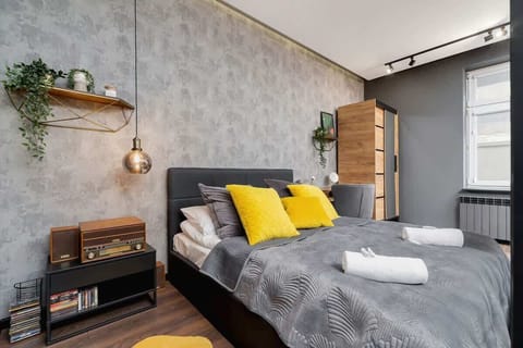 A cozy bedroom with a wallpaper, a neatly made bed with gray bedding, and yellow decorative pillows. A desk and shelves are visible in the corner.