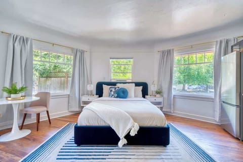 Studio sleeping area with a blue upholstered bed and a small table by the windows.