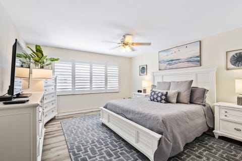 Relax in this bright beach-inspired bedroom featuring a plush king bed, coastal décor, and serene tones. Enjoy natural light through plantation shutters for the perfect seaside retreat.
