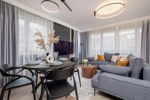 Elegant living room, showing the comfortable seating area, a wall-mounted TV, and floor-to-ceiling windows with light curtains that allow natural light to flood the space.