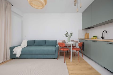 A cozy living room with a teal sofa, wooden flooring, and a stylish open kitchen featuring pastel cabinetry.