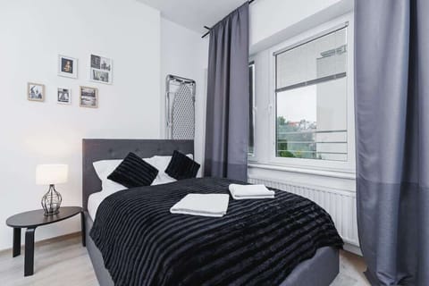A bedroom featuring a neatly made queen-size bed with black bedding. Large windows with gray curtains let in ample natural light.
