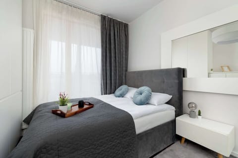 A bedroom featuring double bed with white bedding, decorative pillows, a large window with sheer curtains, and a modern aesthetic.