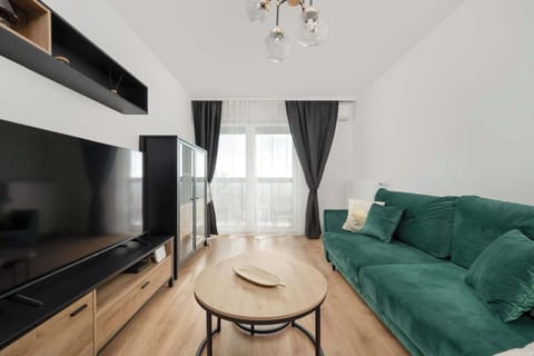Living room with a large green velvet sofa and a round coffee table. On the left, there's a TV stand with a big television. The room is bright and minimalist, with wooden floors and a balcony window with dark curtains.